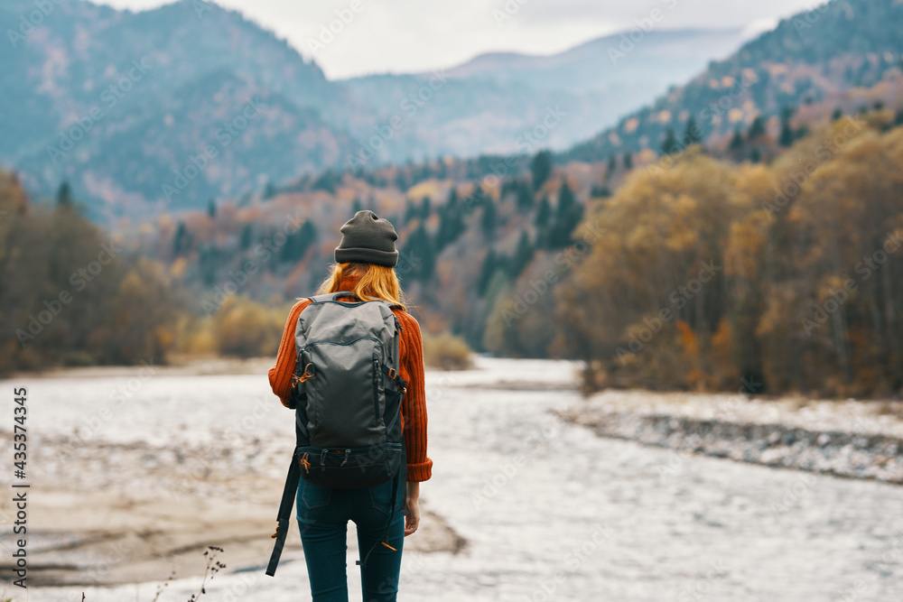 woman in a red sweater cap with a backpack on the river bank and mountains in the distance woman in a red sweater cap with a backpack on the river bank and mountains in the distance
