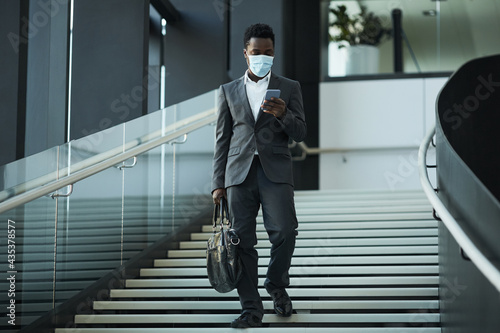 Full length portrait of successful African-American businessman wearing mask and using smartphone while walking towards camera in office building, copy space