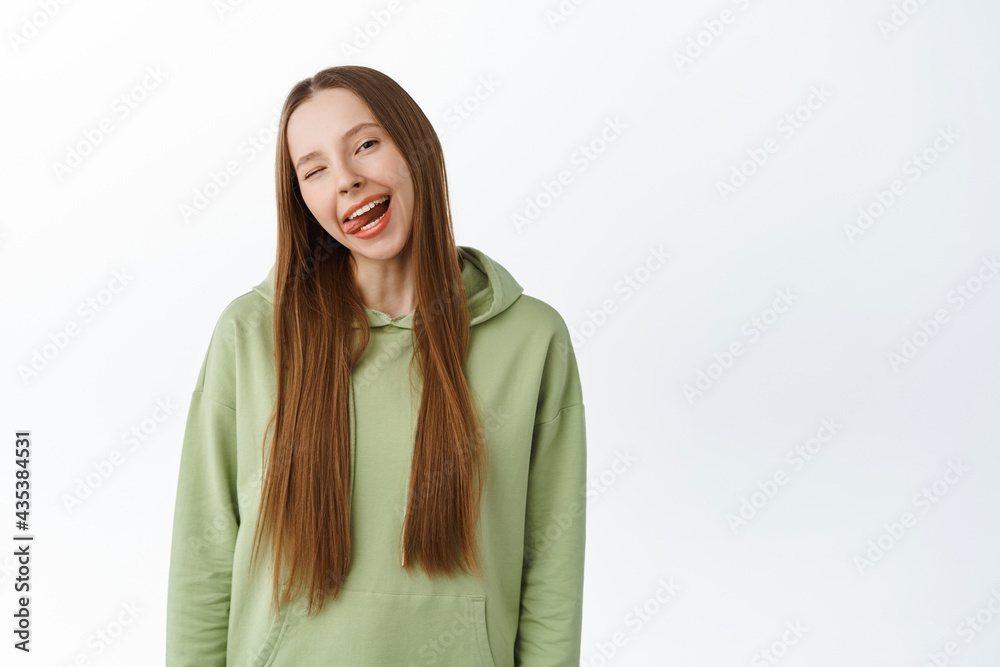 Carefree beautiful millennial girl show tongue, smiling with white perfect teeth and winking, standing in hoodie with positive emotion, studio background Carefree beautiful millennial girl show tongue, smiling with white perfect teeth and winking, standing in hoodie with positive emotion, studio background