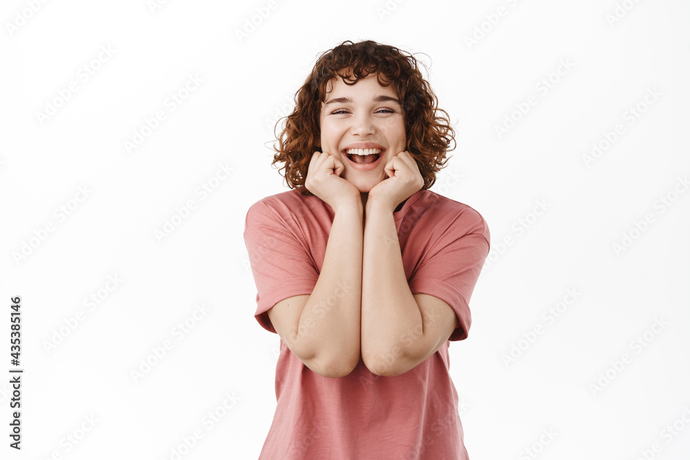 Happy cute curly-haired girl, touching her face with clean natural skin, smiling and laughing at camera, standing in t-shirt against white background Happy cute curly-haired girl, touching her face with clean natural skin, smiling and laughing at camera, standing in t-shirt against white background