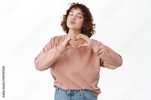 Romantic beautiful girl, waiting for kiss, kissing and showing heart I love you gesture, dreaming of relationship, standing against white background