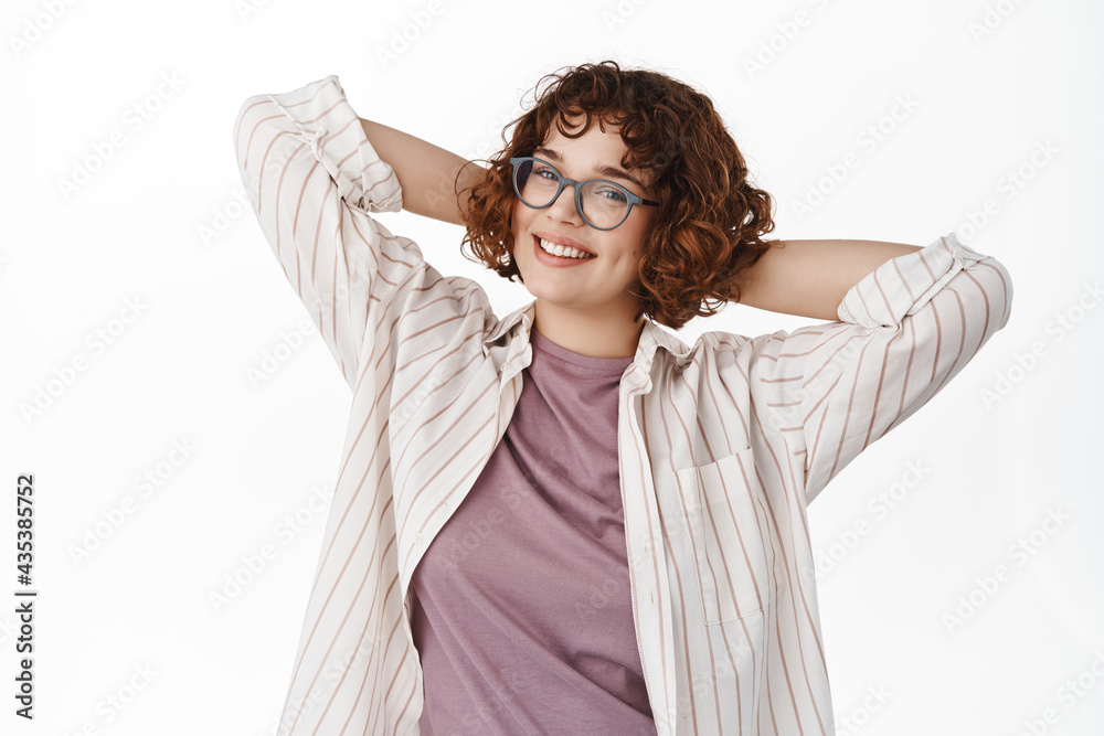 Naklejka premium Portrait of curly girl in glasses resting on day off, relaxing and smiling happy, standing carefree with happy face, looking straight at camera, white background
