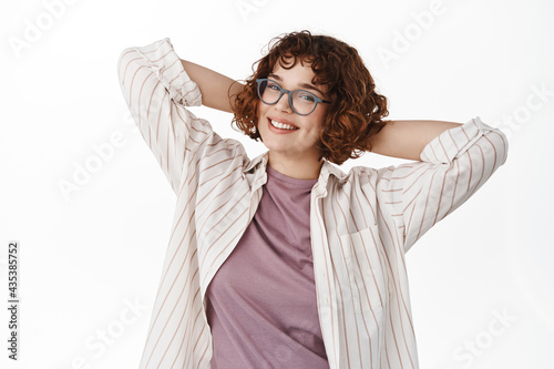 Portrait of curly girl in glasses resting on day off, relaxing and smiling happy, standing carefree with happy face, looking straight at camera, white background