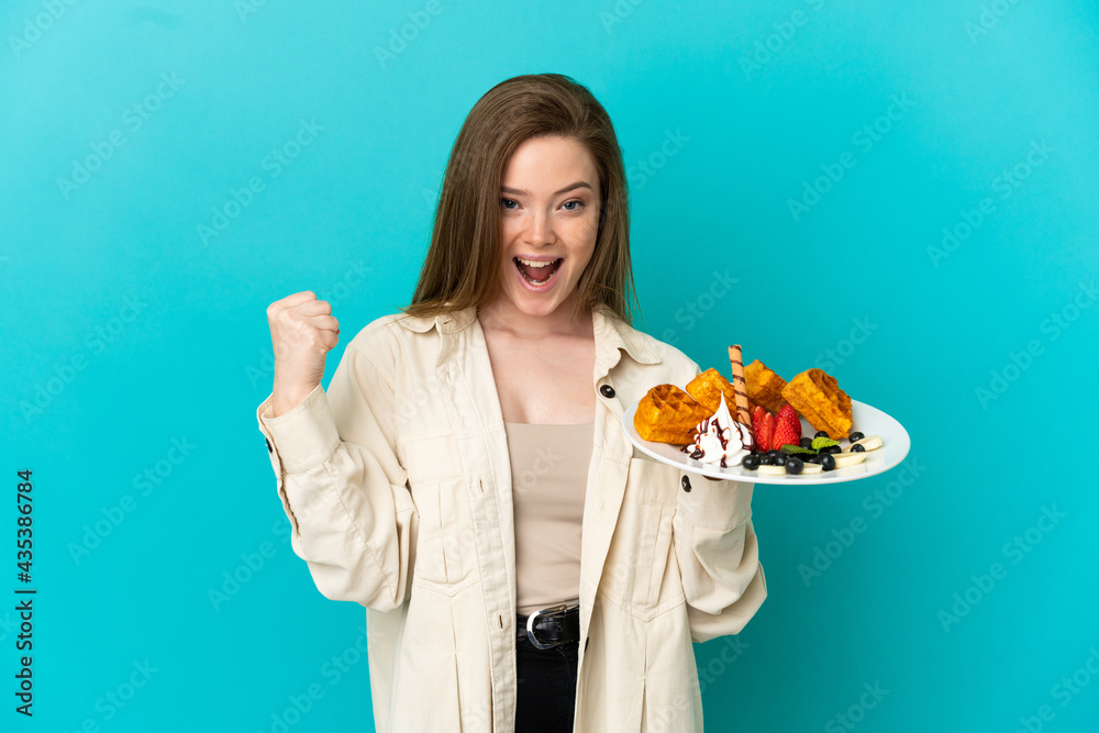 Teenager girl holding waffles over isolated blue background celebrating a victory in winner position Teenager girl holding waffles over isolated blue background celebrating a victory in winner position