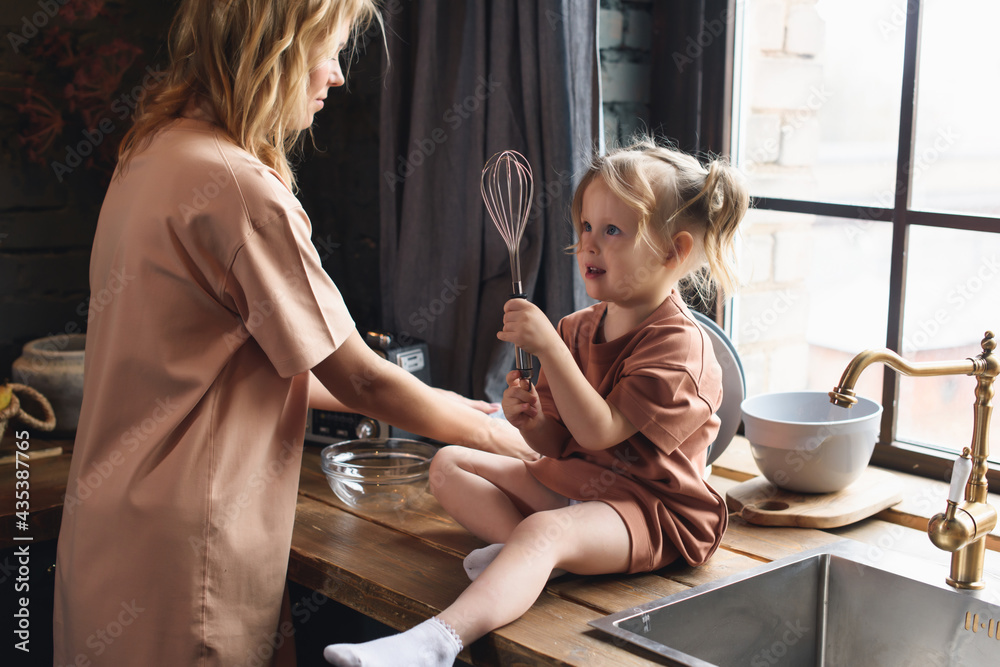 Young mother with her little preschool daughter cook together in the kitchen. Girl beats an egg with a whisk and fooling around