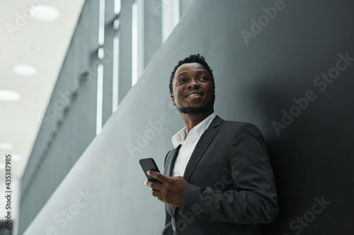 Graphic low angle portrait of carefree African-American businessman holding smartphone while standing against black background in office hall, copy space