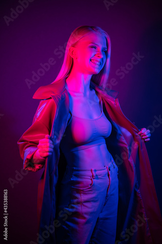 Fashionable model posing in neon light and smiling. Stylishly dressed blonde woman in beautiful clothes.