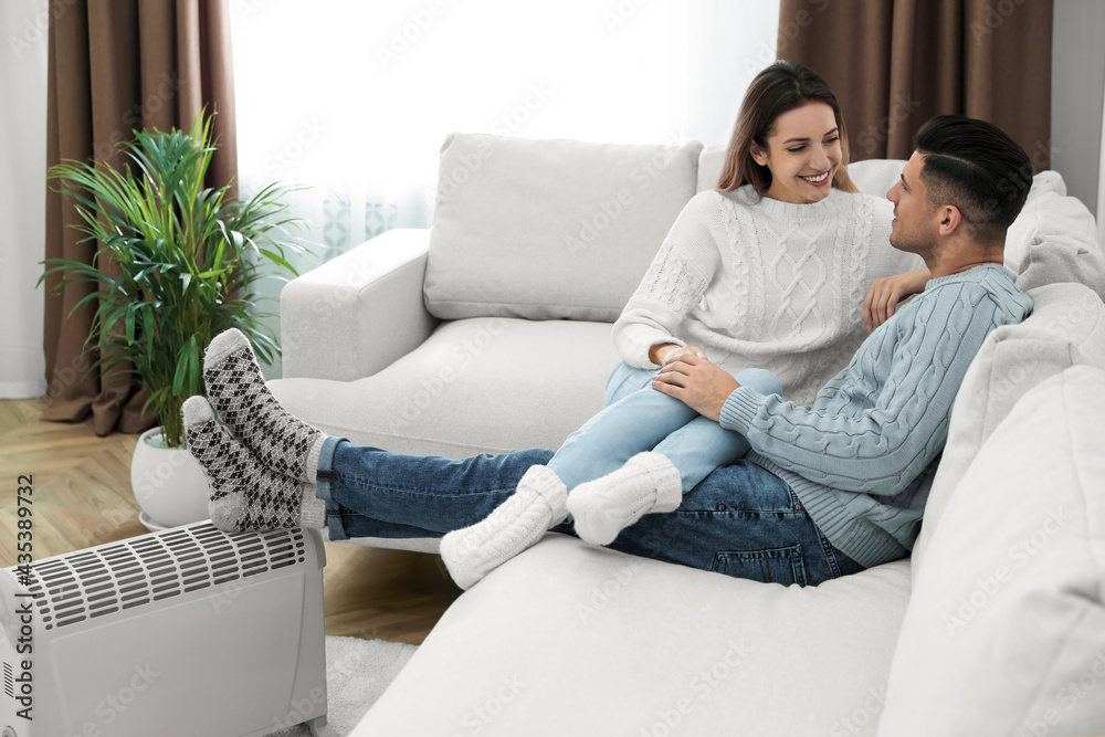 Happy couple sitting on sofa near electric heater at home Happy couple sitting on sofa near electric heater at home