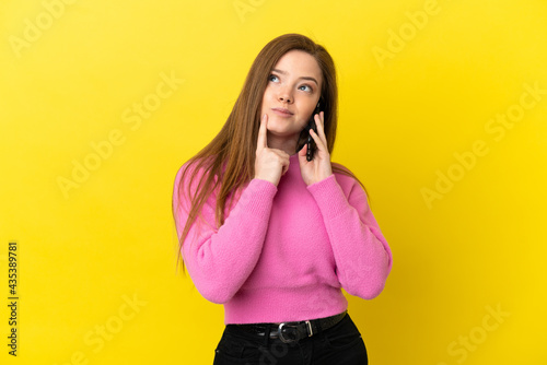 Teenager girl using mobile phone over isolated yellow background having doubts while looking up