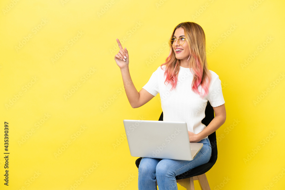 Young woman sitting on a chair with laptop over isolated yellow background pointing up a great idea Young woman sitting on a chair with laptop over isolated yellow background pointing up a great idea