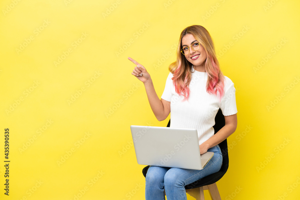 Young woman sitting on a chair with laptop over isolated yellow background pointing to the side to present a product Young woman sitting on a chair with laptop over isolated yellow background pointing to the side to present a product