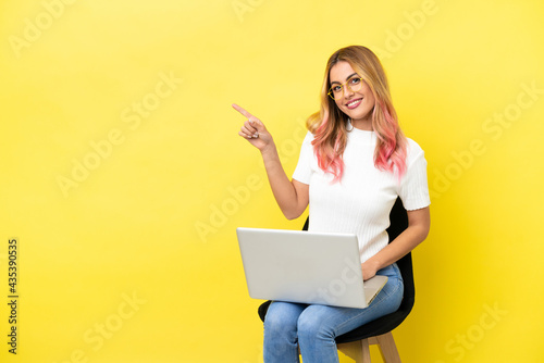 Young woman sitting on a chair with laptop over isolated yellow background pointing to the side to present a product