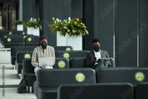 Graphic wide angle portrait of African-American businessman wearing mask while working with laptop in waiting lounge at airport with social distancing