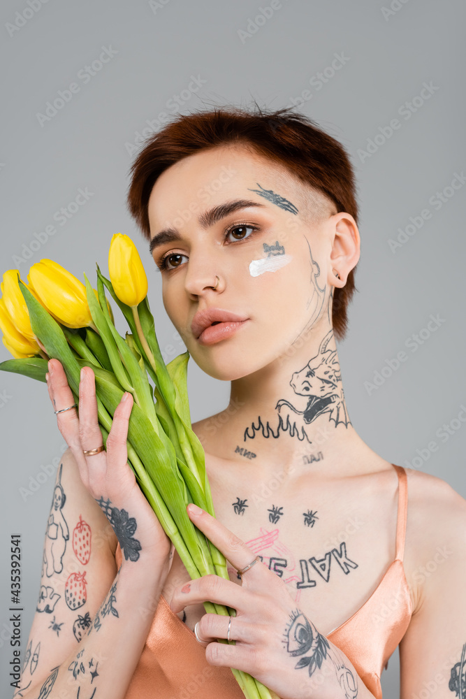 Naklejka premium tattooed young woman with cosmetic cream on face holding tulips isolated on grey