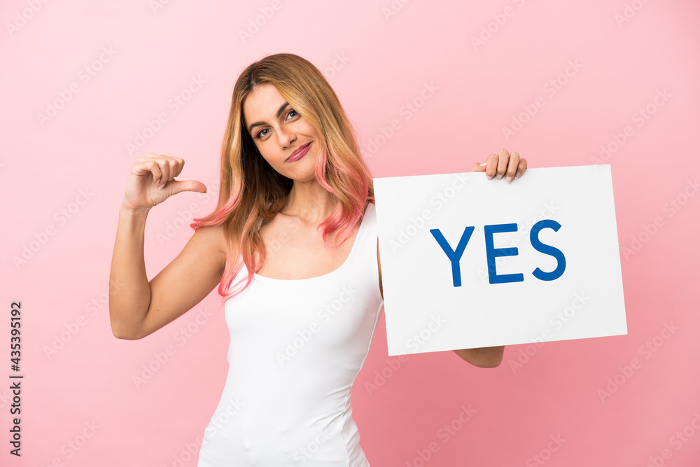 Young woman over isolated pink background holding a placard with text YES with proud gesture Young woman over isolated pink background holding a placard with text YES with proud gesture