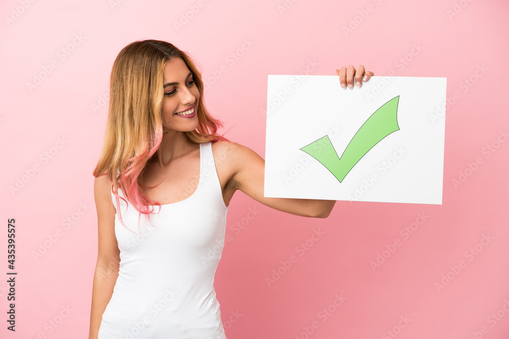 Young woman over isolated pink background holding a placard with text Green check mark icon with happy expression Young woman over isolated pink background holding a placard with text Green check mark icon with happy expression