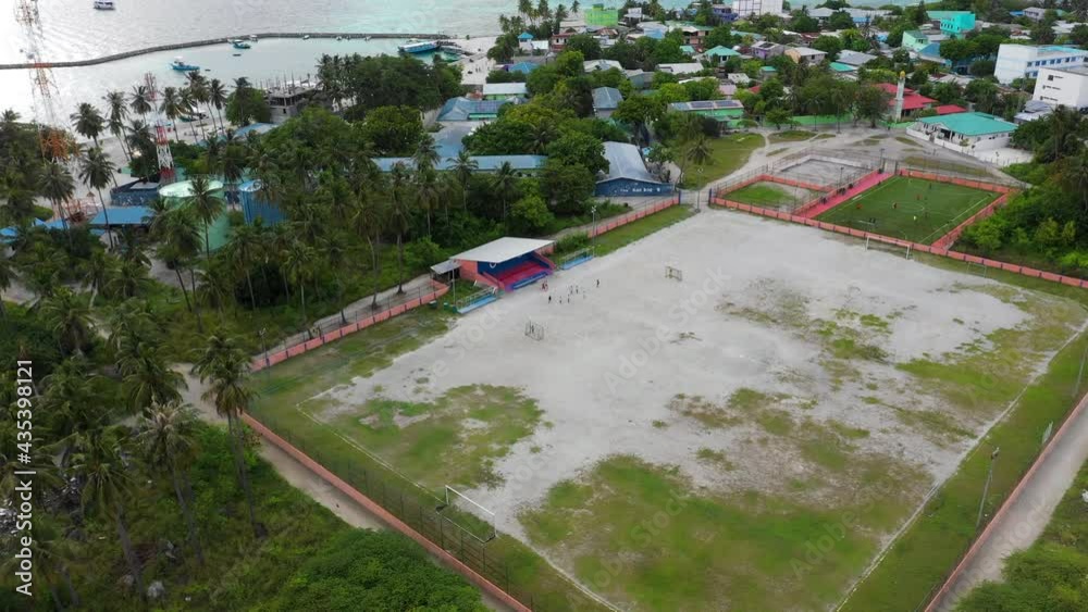Aerial Moving Forward And Panning A Soccer Field With A Small Group Of Players On A Tropical Island, With Palm Trees, A Sandy Field, And The Rooftops Of Nearby Buildings - Guraidhoo, Maldives Aerial Moving Forward And Panning A Soccer Field With A Small Group Of Players On A Tropical Island, With Palm Trees, A Sandy Field, And The Rooftops Of Nearby Buildings - Guraidhoo, Maldives