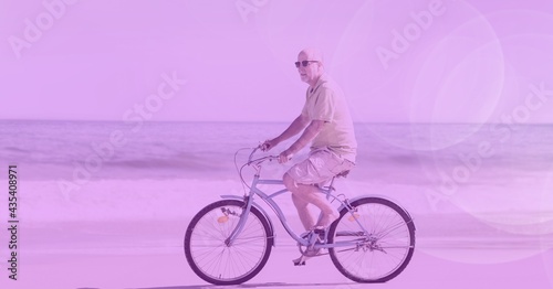 Composition of senior man riding bicycle on beach by sea with purple tint