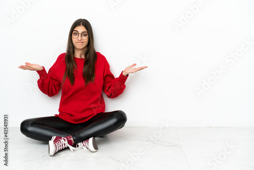 Young woman sitting on the floor isolated on white background having doubts while raising hands