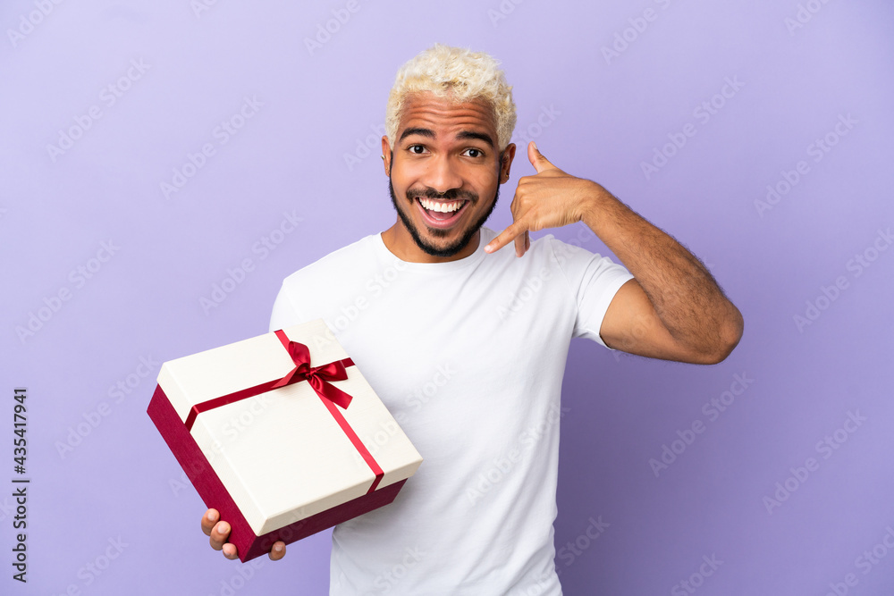 Young Colombian man holding a gift isolated on purple background making phone gesture. Call me back sign Young Colombian man holding a gift isolated on purple background making phone gesture. Call me back sign