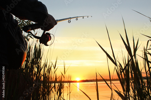 Close-up of a fishing rod with a reel in male hands against the backdrop of the sunset on the river in the wild. Evening fishing with a rod on the river. Country rest, camping