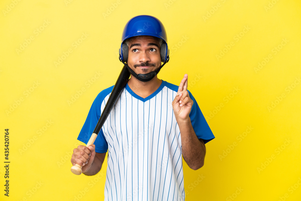 Young Colombian latin man playing baseball isolated on yellow background with fingers crossing and wishing the best