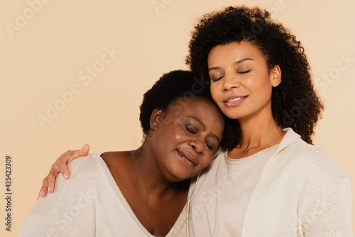 calm african american adult daughter and middle aged mother hugging with closed eyes isolated on beige