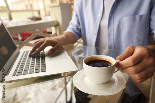 Man with cup of coffee working on laptop at outdoor cafe in morning, closeup