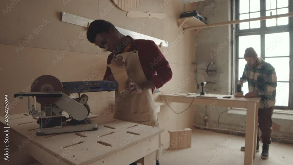 Medium shot of African-American male carpenter standing at workbench and sanding board edges using grinding machine while his male Caucasian colleague working on background