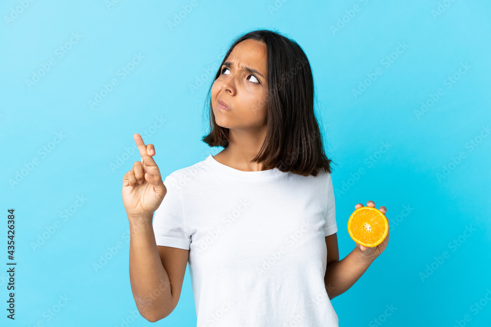 Young asian girl holding an orange isolated on blue background with fingers crossing and wishing the best Young asian girl holding an orange isolated on blue background with fingers crossing and wishing the best