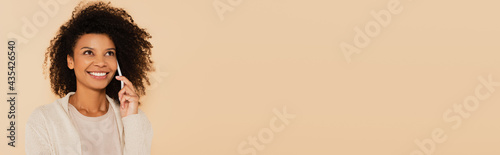 happy african american woman speaking on cellphone and looking up isolated on beige, banner