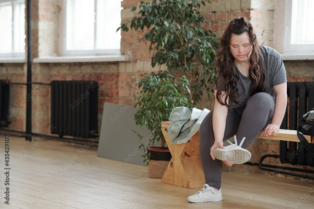 Young woman with Down syndrome putting on sports-shoes before yoga training in gym