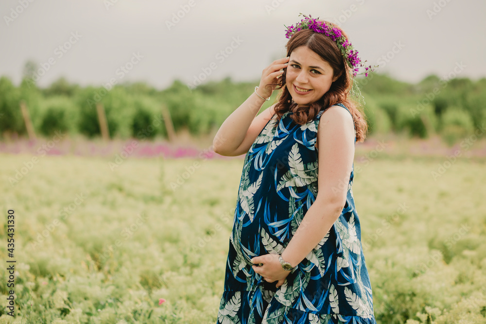 Close up of young smiling pregnant woman in wreath of flowers enjoying maternity standing in field and looking at camera Close up of young smiling pregnant woman in wreath of flowers enjoying maternity standing in field and looking at camera