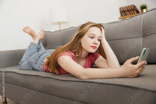 Relaxed smiling young young woman texting on cellphone lying on sofa at home,