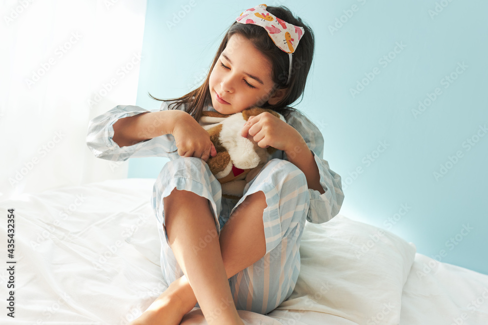 Playful little girl in pajama playing with a toy at bed in the modern cozy sunny bedroom. Cute kid resting on the bed in the morning at home during her birthday. Playful little girl in pajama playing with a toy at bed in the modern cozy sunny bedroom. Cute kid resting on the bed in the morning at home during her birthday.