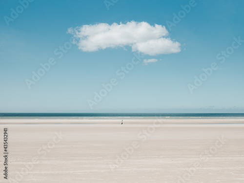 Fototapeta Naklejka Na Ścianę i Meble -  Person walking alone far away on a beach with blue sky and a single cloud
