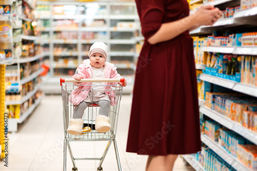 Shopping. A toddler sits in a grocery cart and looks around while his mother picks out groceries. Family shopping in the supermarket