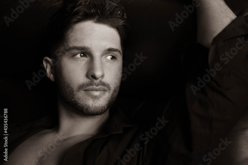 Portrait of attractive young man with beard, open shirt revealing chest, sitting in leather armchair, looking at camera