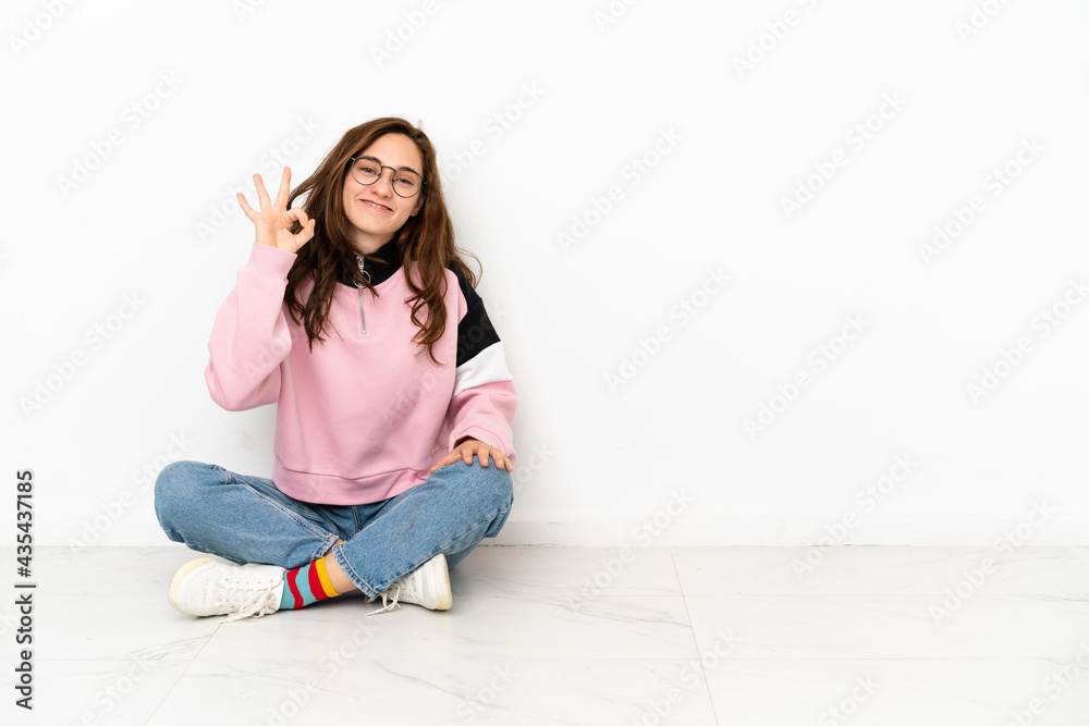 Young caucasian woman sitting on the floor isolated on white background showing ok sign with fingers Young caucasian woman sitting on the floor isolated on white background showing ok sign with fingers