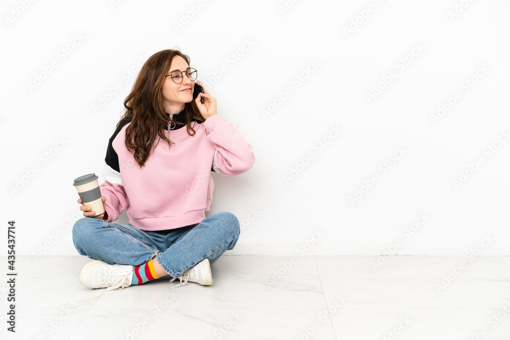 Young caucasian woman sitting on the floor isolated on white background holding coffee to take away and a mobile Young caucasian woman sitting on the floor isolated on white background holding coffee to take away and a mobile