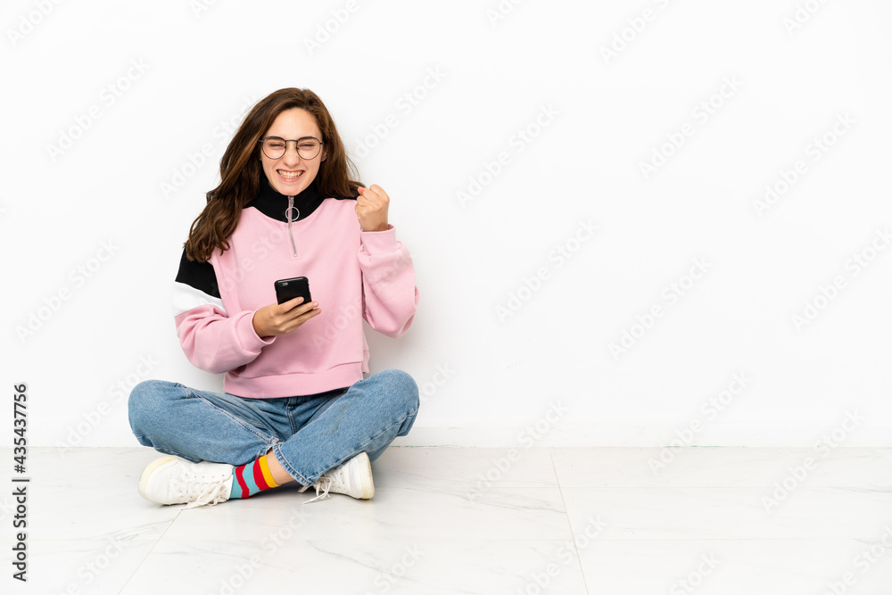 Young caucasian woman sitting on the floor isolated on white background with phone in victory position