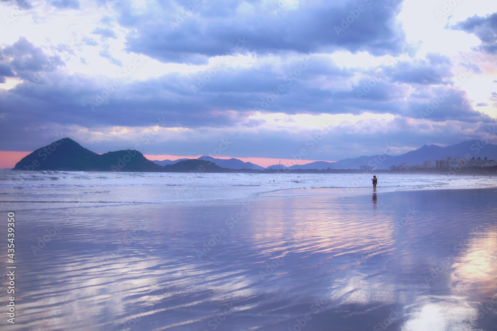 
A man alone far away on the beach sand. Afternoon with bluish skies, lots of clouds and calm waves. Mountains in the background and cloudy blue sky