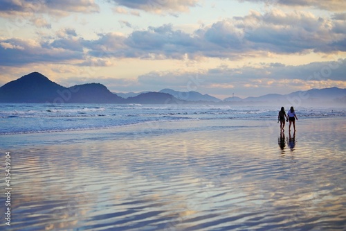 Couple of women walking on the beach sand. Calm sea with few waves. Diversity, human love. Mountains in the background and cloudy blue sky