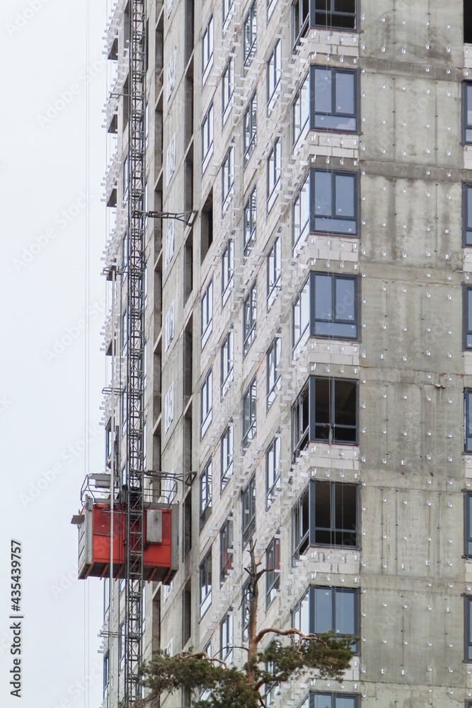 Red construction lift on facade of residential building under construction. Elevator is used to lift workers and building materials outside house Red construction lift on facade of residential building under construction. Elevator is used to lift workers and building materials outside house