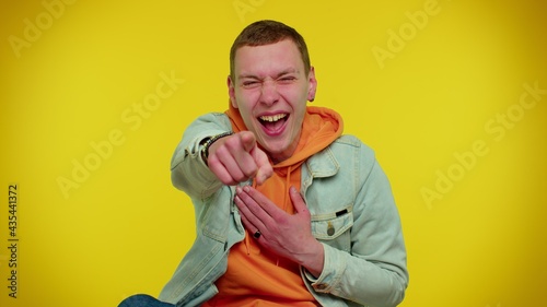 Amused teen stylish man in denim jacket pointing finger to camera, laughing out loud, taunting making fun of ridiculous appearance, funny joke. Young guy boy posing on yellow studio wall background photo