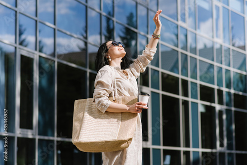 Happy brunette young woman with sunglasses and bag holding coffee walking in the city. Lifestyle portrait of smiling woman