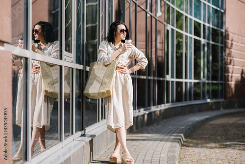 Happy brunette young woman with sunglasses and bag holding coffee walking in the city. Lifestyle portrait of smiling woman