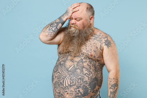 Foto Side view sad young fat pudge obese chubby overweight blue-eyed bearded man 30s has big belly with naked tattooed torso put hand on face facepalm epic fail gesture isolated on pastel blue background