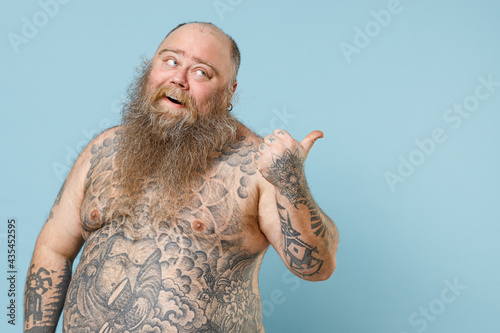 Young fun fat pudge obese chubby overweight blue-eyed bearded man 30s has big belly with naked tattooed torso point thumb aside back worksace area isolated on pastel blue background studio portrait
