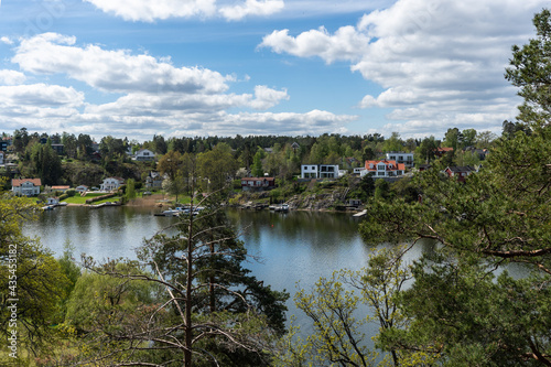 Panoramic view of the Baltic Sea Bay on sunny spring day. Rocky shores of Scandinavia covered with evergreen forests. Traditional Swedish red wooden houses cabins on the coast. Blue sky white clouds.